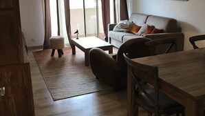 A living room with a sofa, coffee table, and dining table. There is a window with curtains, a hanging light fixture, and a wall-mounted artwork.
