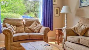 A living room with a beige sofa, a wooden coffee table, and a side table with a lamp. There are two windows with blue curtains, and a framed picture on the wall. - Craik Forestry Cottage (Craik, near Hawick)