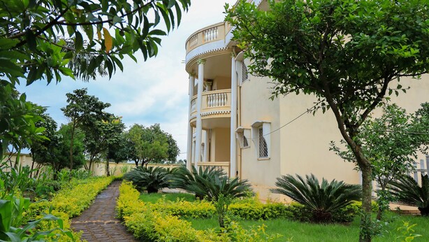 A building with a curved balcony, surrounded by lush greenery and a paved pathway. -  Manoir Des Innoncents (Bafoussam)