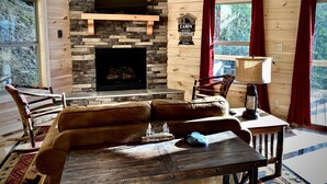 Interior acogedor de una cabaña con chimenea de piedra, paredes de madera, un sofá de cuero y un televisor sobre la chimenea.