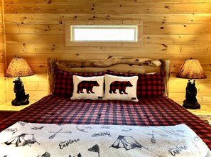 A cozy cabin room with a wooden headboard, checkered bedding, two lamps, and a window above the bed.