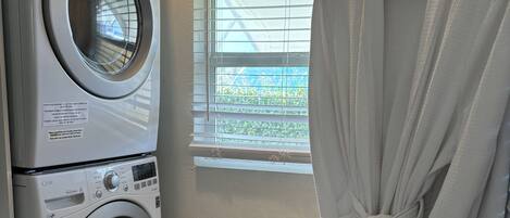 Un área de lavandería con lavadora y secadora de carga frontal, una ventana con persianas y una bata blanca colgada en un perchero.