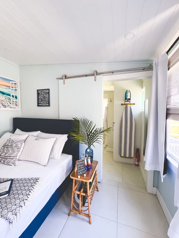 Un dormitorio con cama, una mesita de noche con una planta, una pared con un cartel enmarcado y vistas a un pasillo.