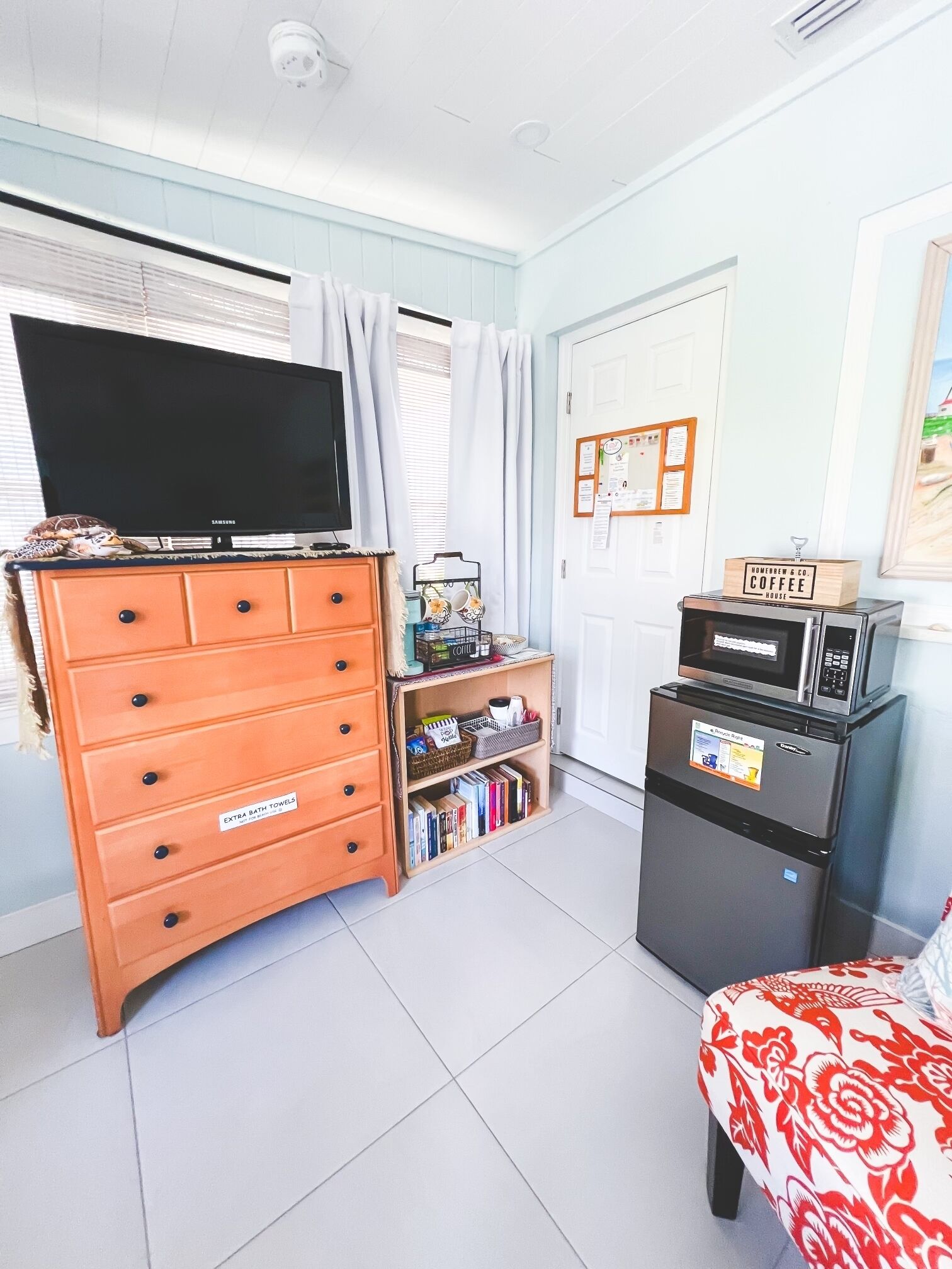 A room with a television on a dresser, a refrigerator, a bookshelf, and a bed with a floral bedspread.