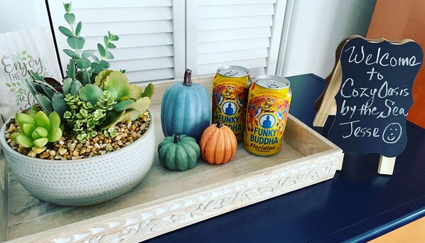 A tray with a potted plant, two pumpkins, and two cans of drink on a wooden surface. A chalkboard sign reads "Welcome to Cozy Oasis by the Sea Jesse".