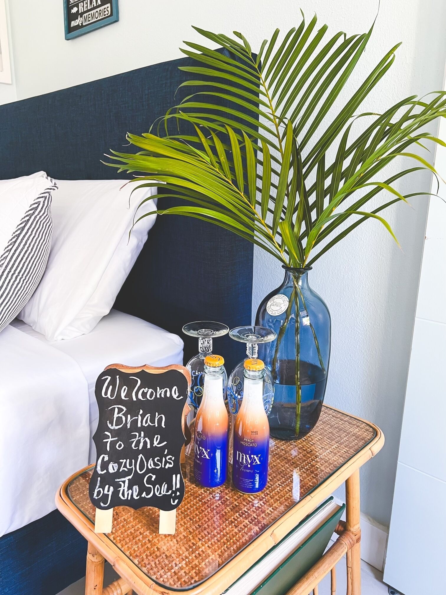 A cozy room with a bed, a wicker table with a welcome sign, a potted plant, and bottles of beverages.