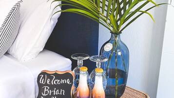 A cozy room with a bed, a wicker table with a welcome sign, a potted plant, and bottles of beverages.