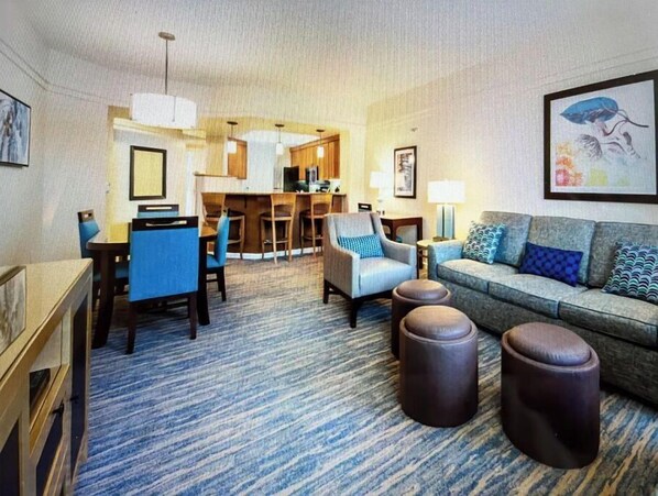 Ein Hotelzimmer mit Wohnbereich, ausgestattet mit einer Couch, Hocker und einem Essbereich mit Stühlen und Tisch.