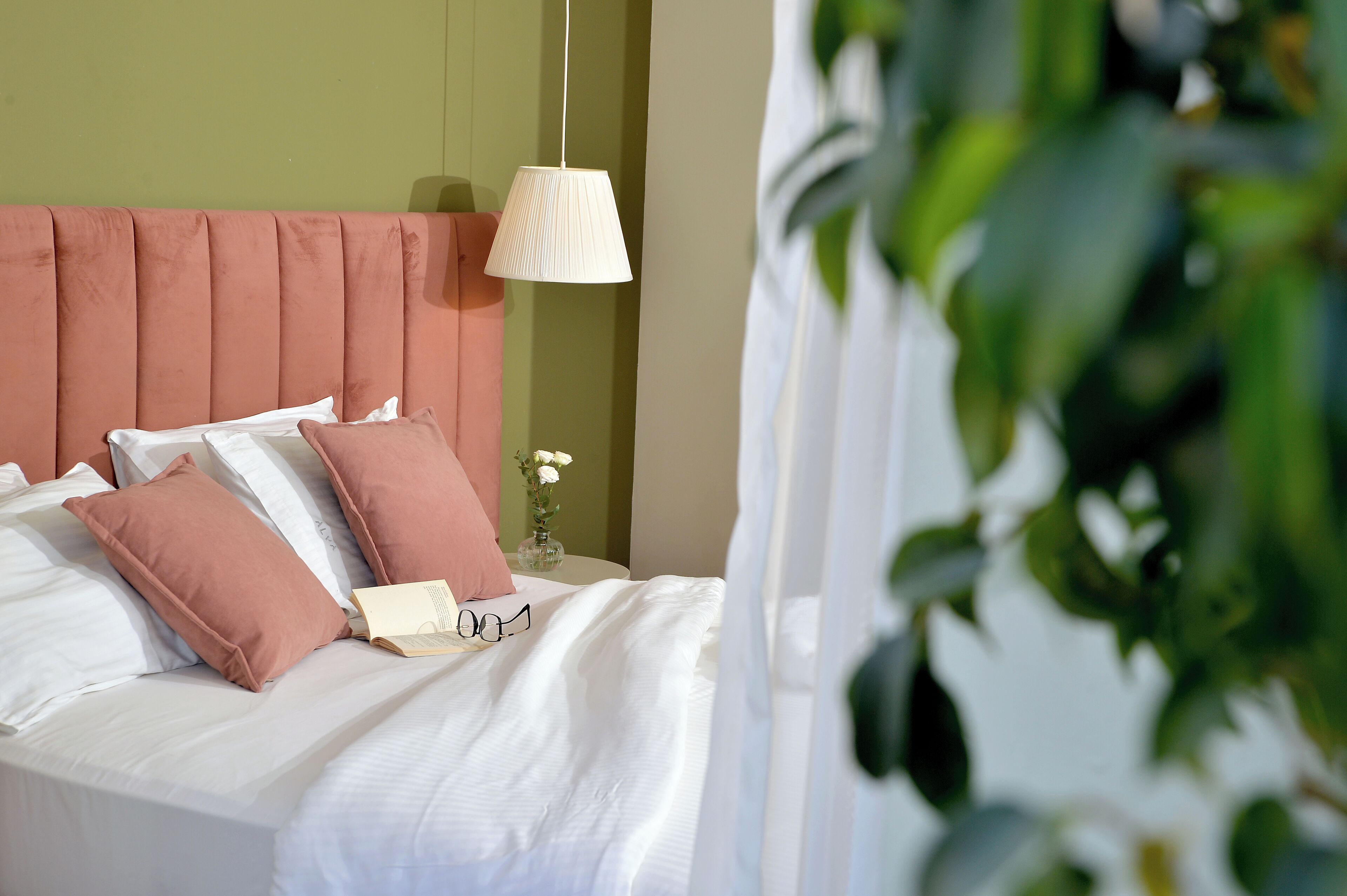 A neatly made bed with white and pink pillows, a white lamp, and a vase with flowers on a green wall.