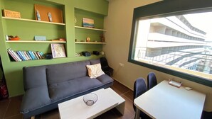 A modern living room with a grey sofa, a white coffee table, and a dining area with a white table and two chairs. There are shelves with books and decorative items, and a window with a view of a building.