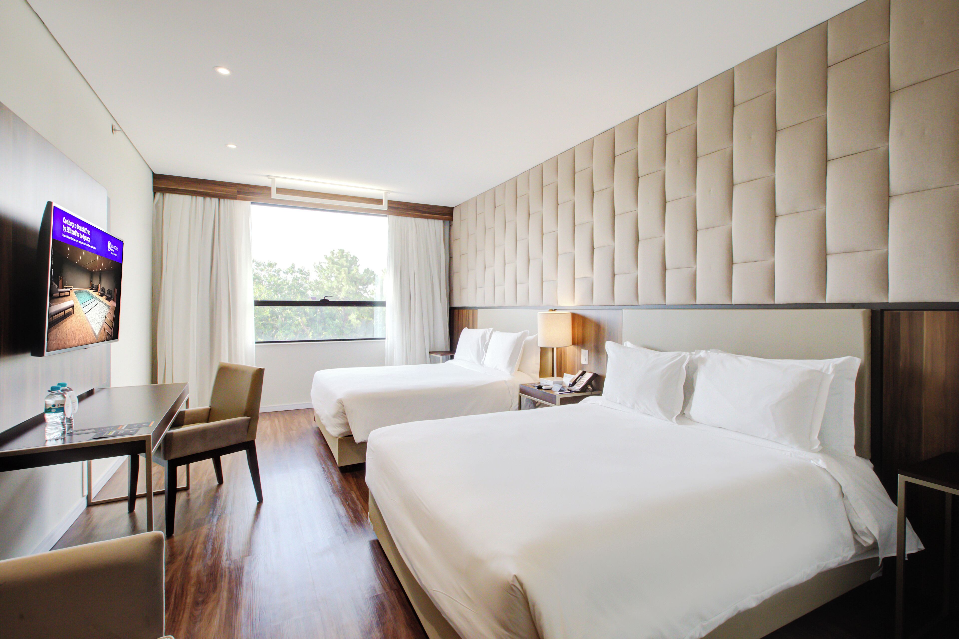 A hotel room with two beds, a desk, a chair, a TV, and a window with curtains.