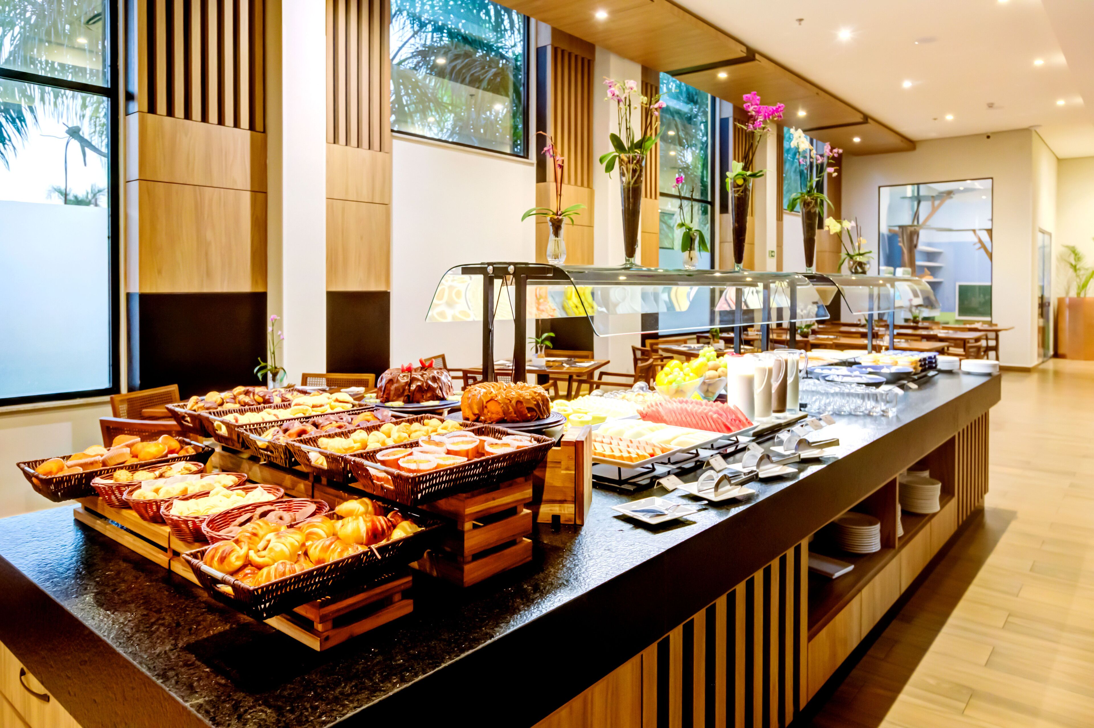 A buffet with various food items, including pastries and fruits, in a modern hotel setting with wooden accents and large windows.