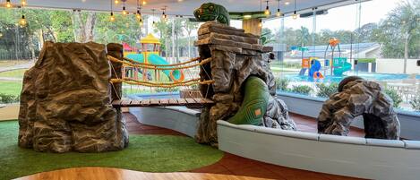 A play area with rock structures, a rope bridge, and a view of a swimming pool and playground.