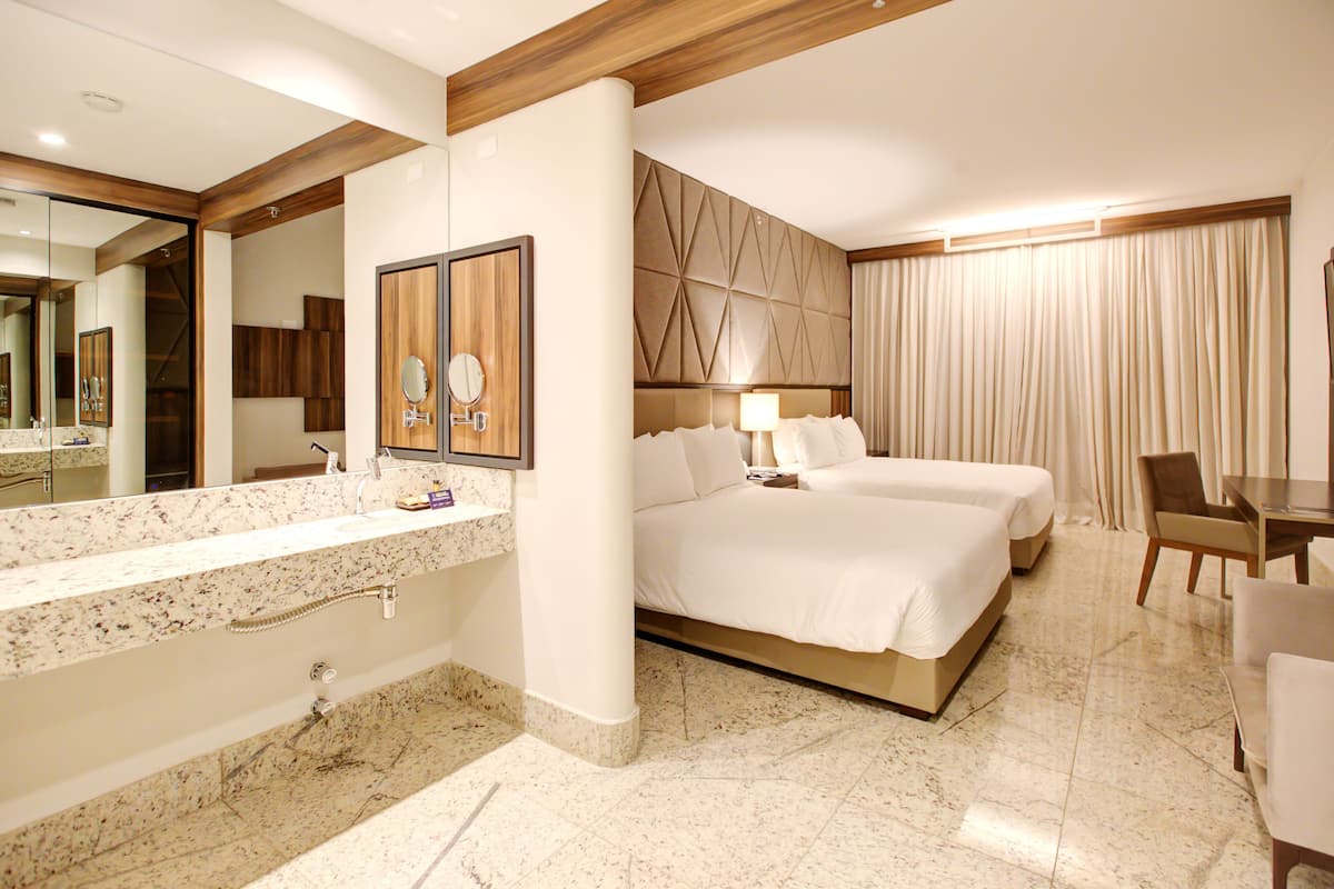 A hotel room with a marble bathroom, two beds, a dining table, and a large mirror.