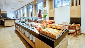 Uma área de buffet com diversas opções de comida, um espaço de estar com mesas e cadeiras, e uma grande janela que proporciona iluminação natural.