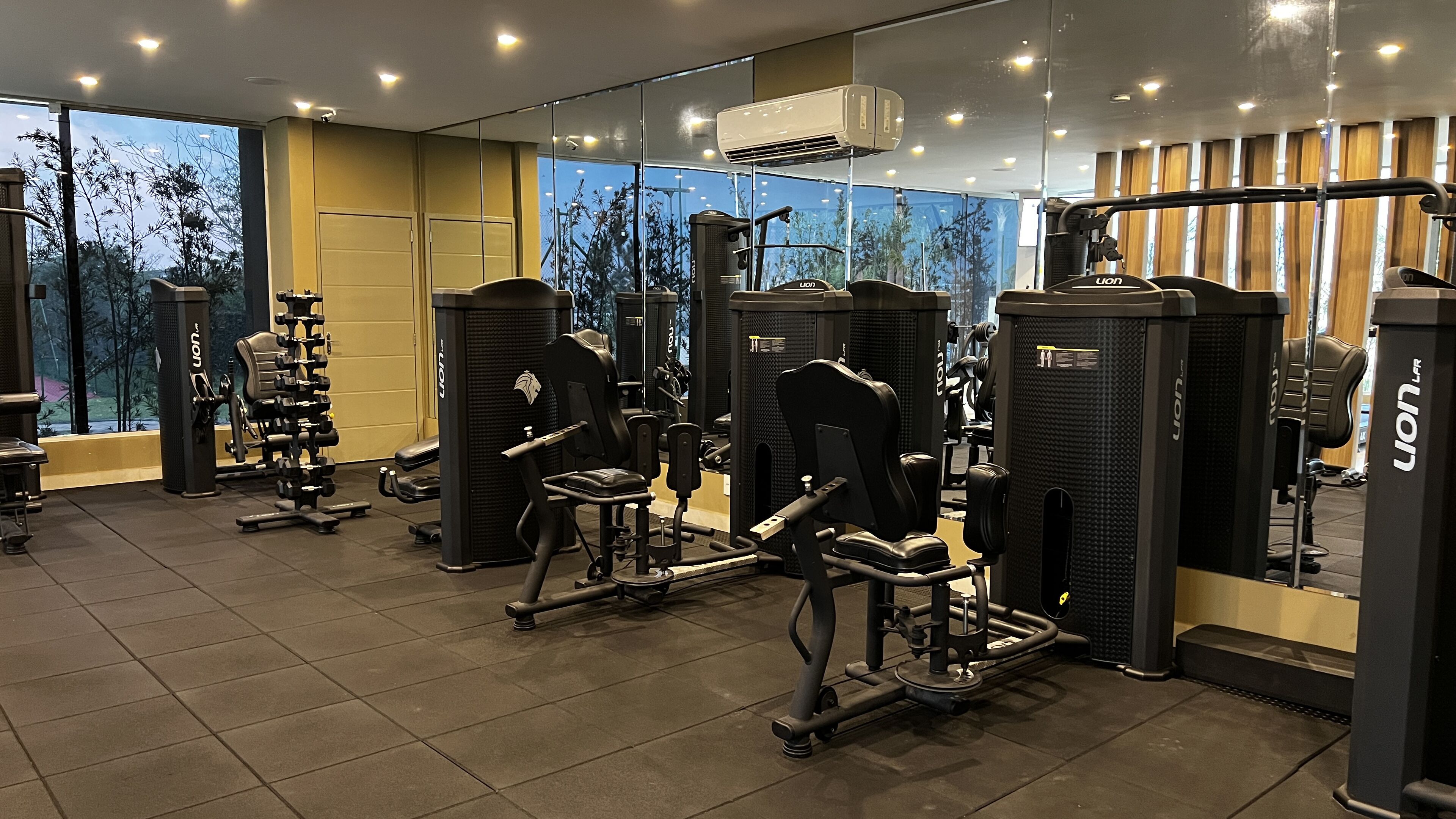 A gym with various exercise equipment, large windows with a view of trees, and an air conditioning unit on the ceiling.