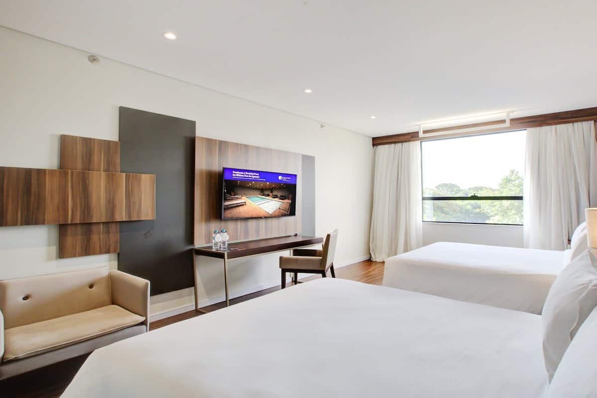 A hotel room with two beds, a desk with a TV, and a view of the outdoors.