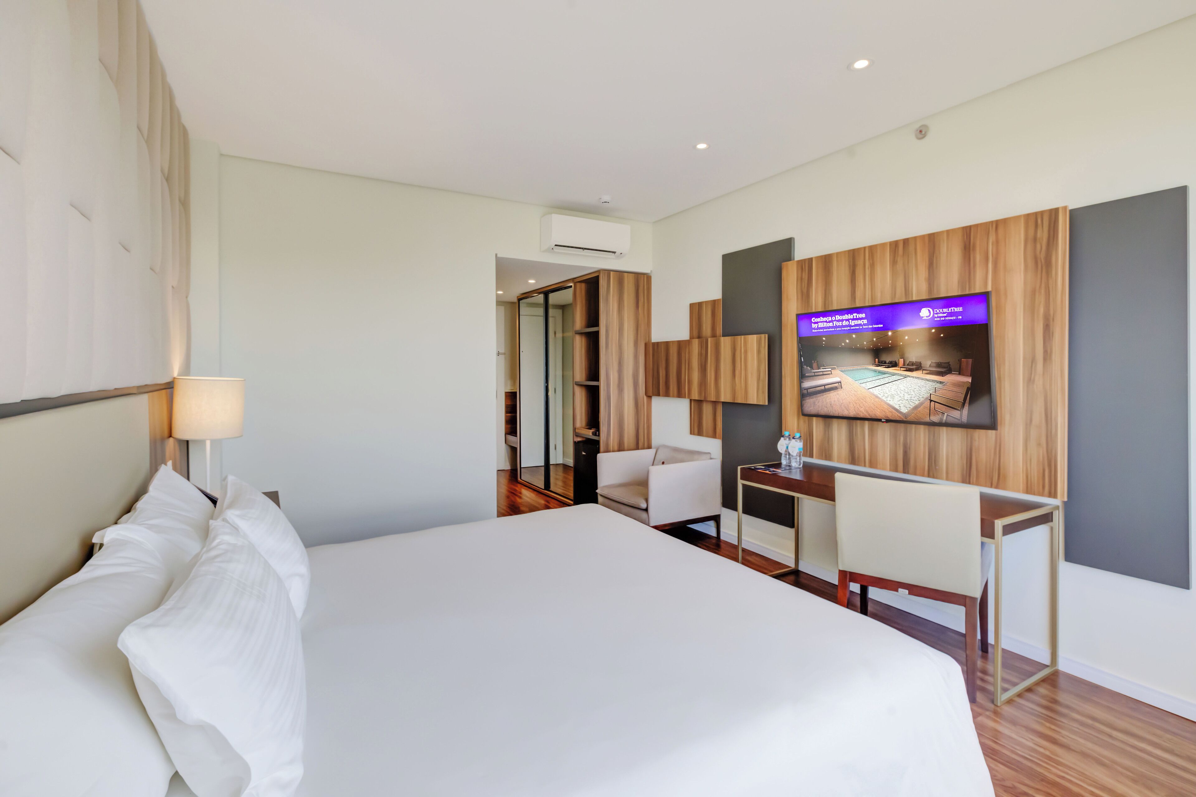 A modern hotel room with a large bed, a desk, a chair, a television mounted on the wall, and a mirror.