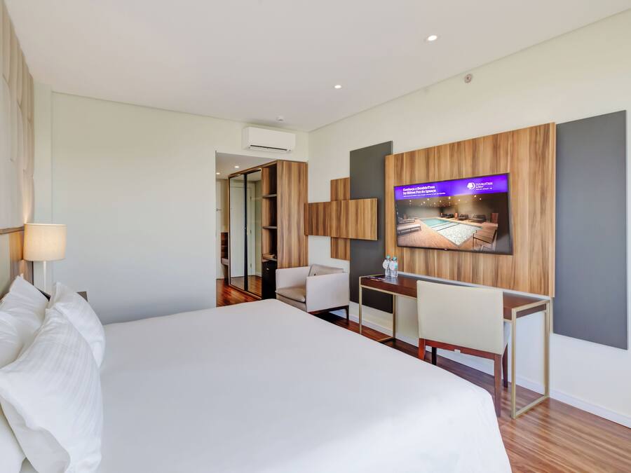 A modern hotel room with a large bed, a desk, a chair, a television mounted on the wall, and a mirror.