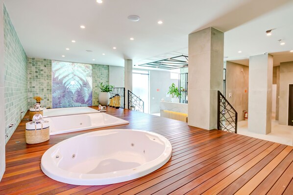 Un baño espacioso con dos tinas grandes, piso de madera y pared revestida de azulejos verdes.