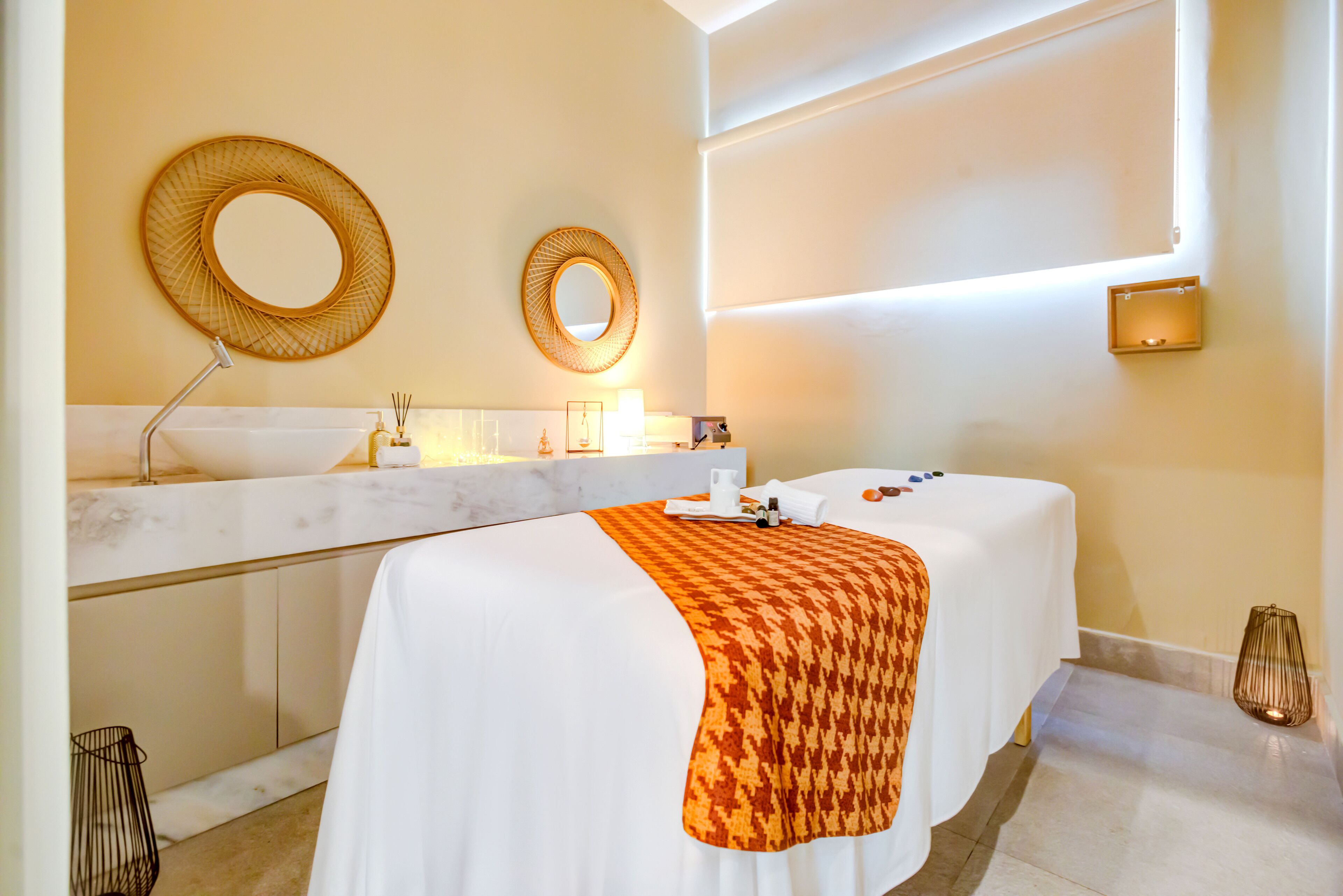 A hotel room with a large white bathtub, two round mirrors, a white bed with an orange blanket, and a marble countertop with toiletries.
