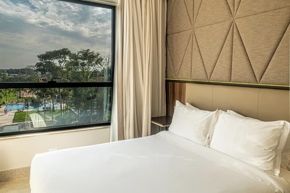 A hotel room with a large window, a bed with white linens, and a tufted headboard.
