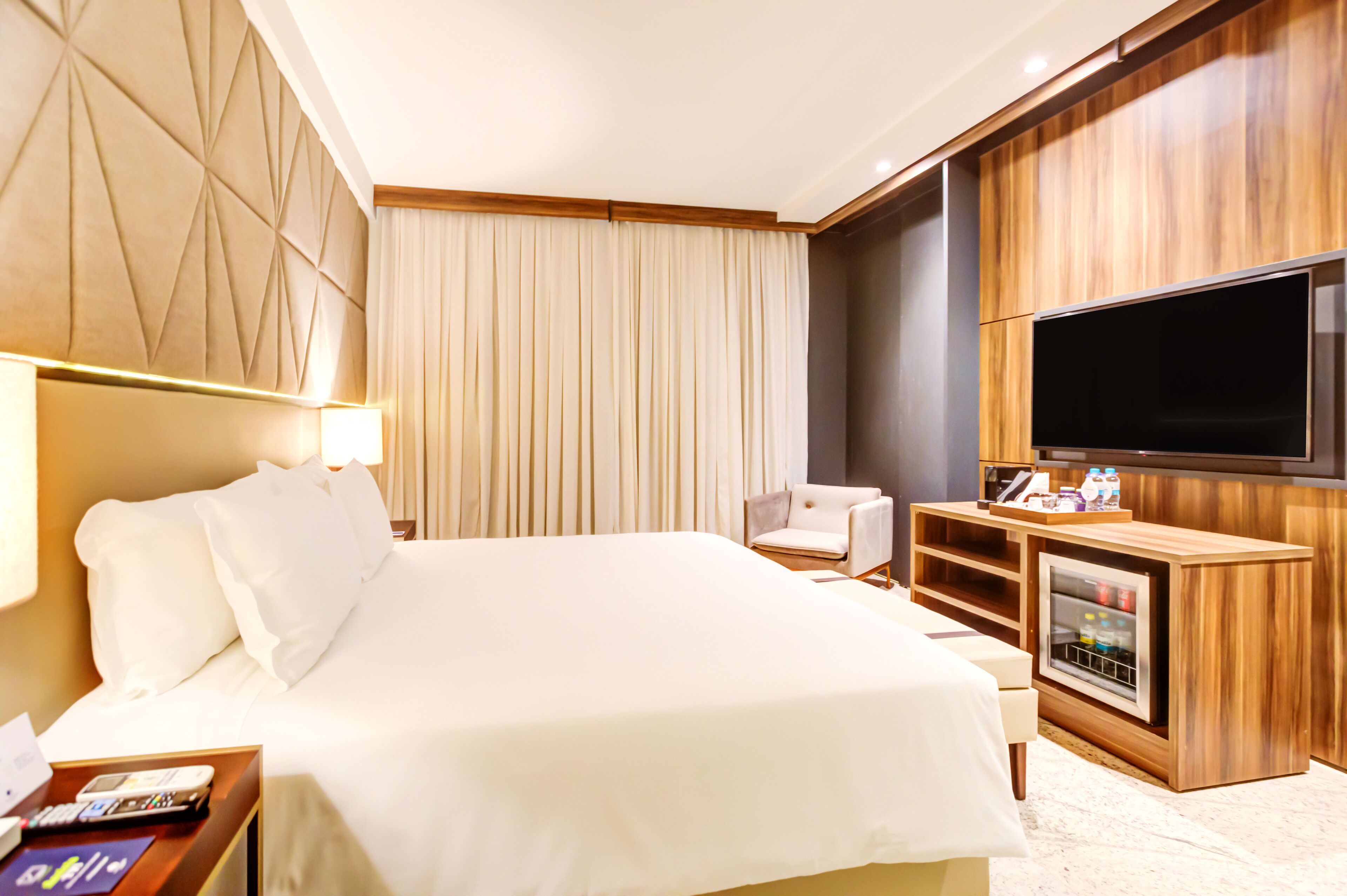 A hotel room with a large bed, a flat-screen TV, a seating area with a chair, and a minibar.