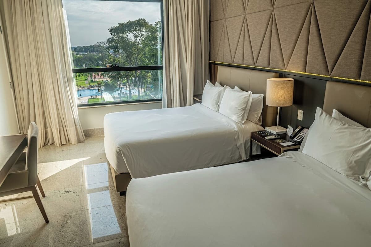 A hotel room with two beds, a desk, and a window with a view of a pool and trees.
