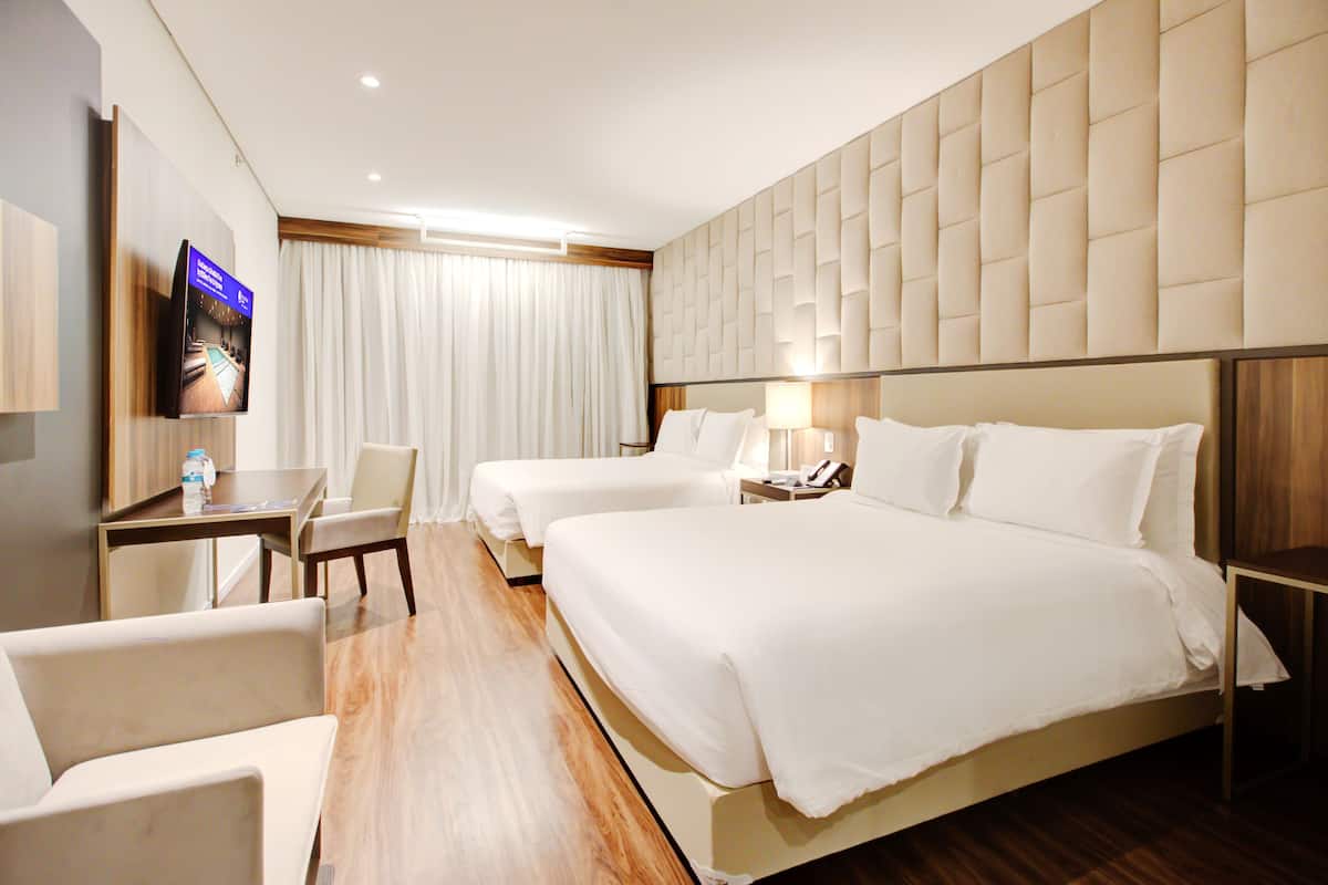 A hotel room with a large bed, two smaller beds, a desk with a chair, a TV, and a bathroom with a mirror and toilet.