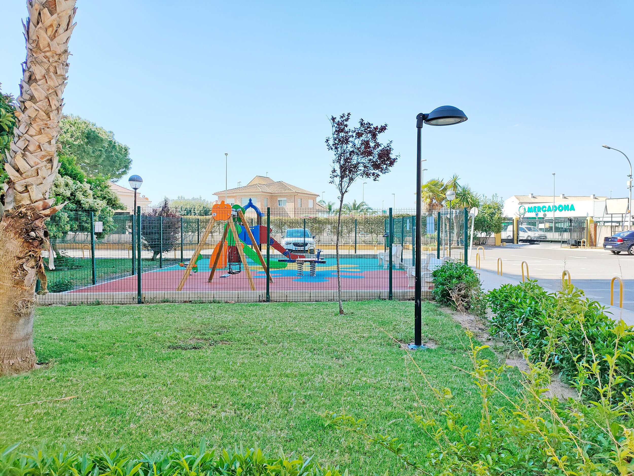 A playground with a slide and swings, surrounded by a fence and greenery.