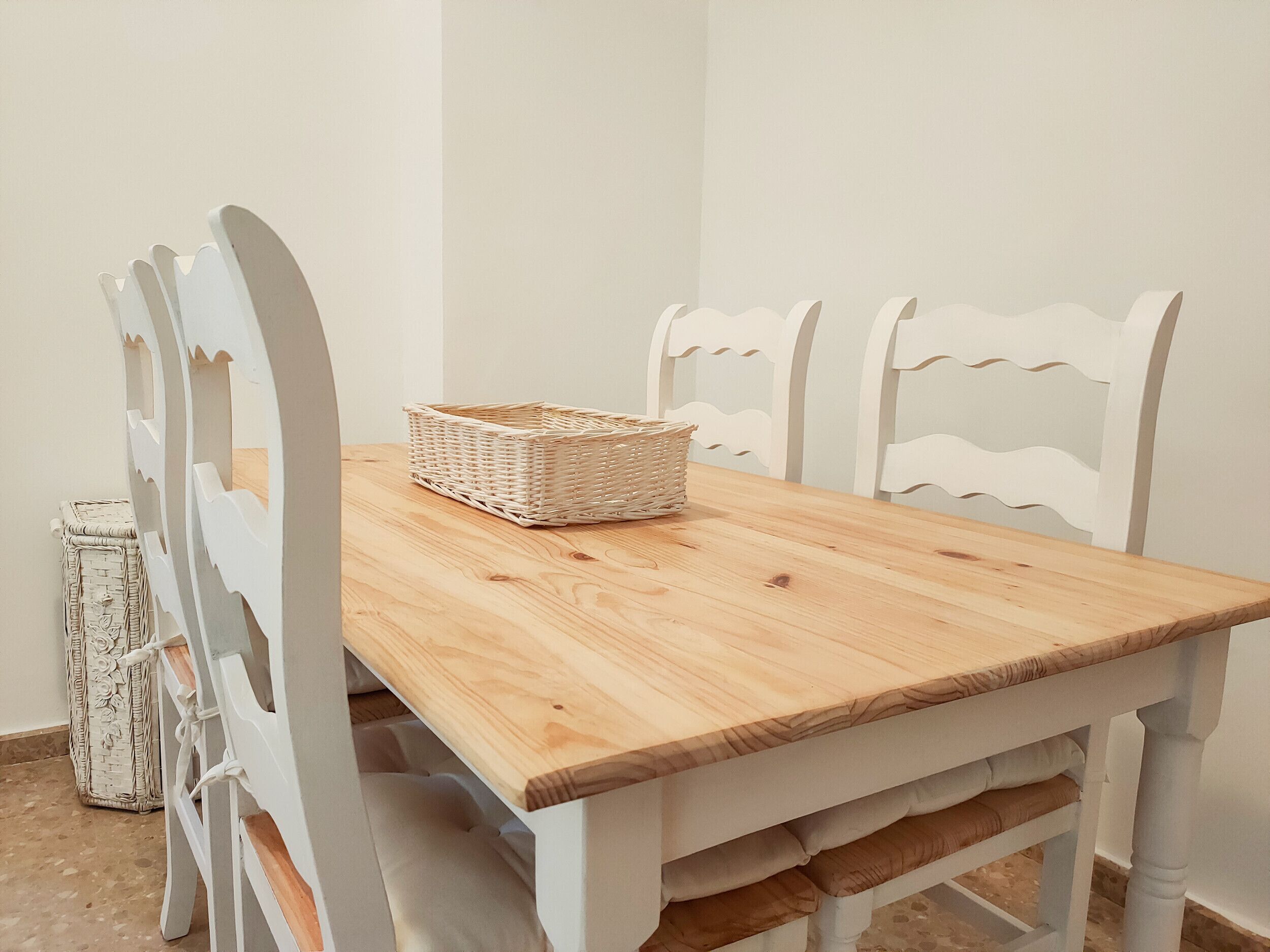 Una mesa de comedor de madera con sillas blancas, una cesta tejida y un adorno de mimbre.