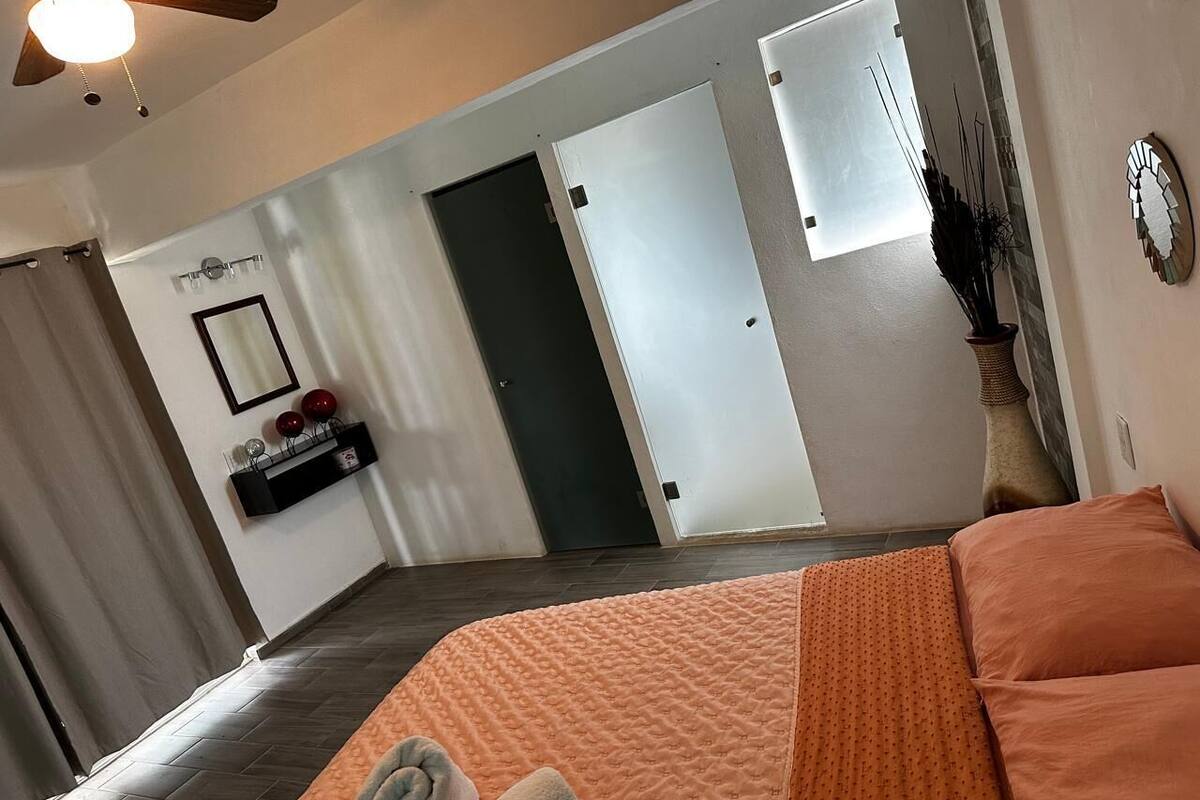 Une chambre à coucher comprenant un lit, un ventilateur de plafond, un miroir, une étagère avec des objets décoratifs et une porte donnant sur une autre pièce.
