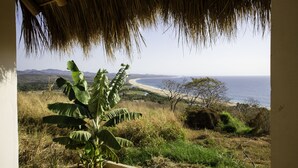 Vista desde una construcción con techo de paja que da a una playa y montañas lejanas.