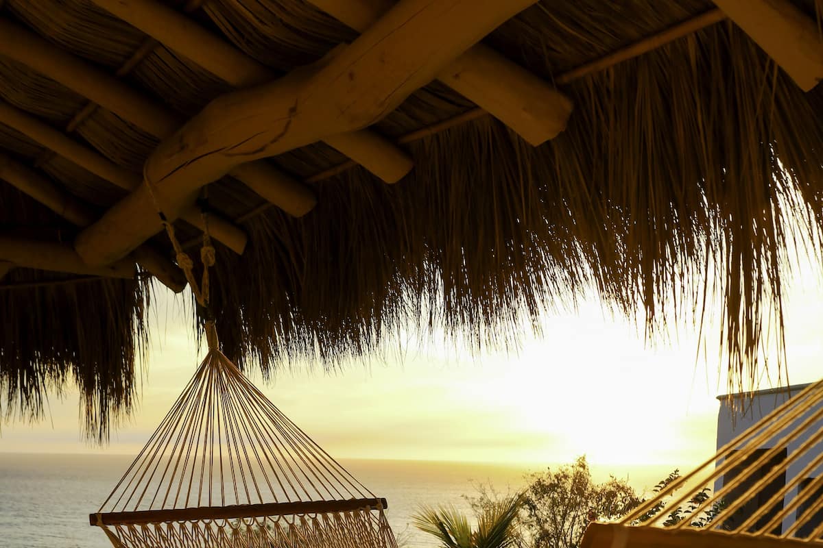 Un hamac suspendu sous un toit de chaume, avec vue sur une plage bordée de palmiers et un coucher de soleil.
