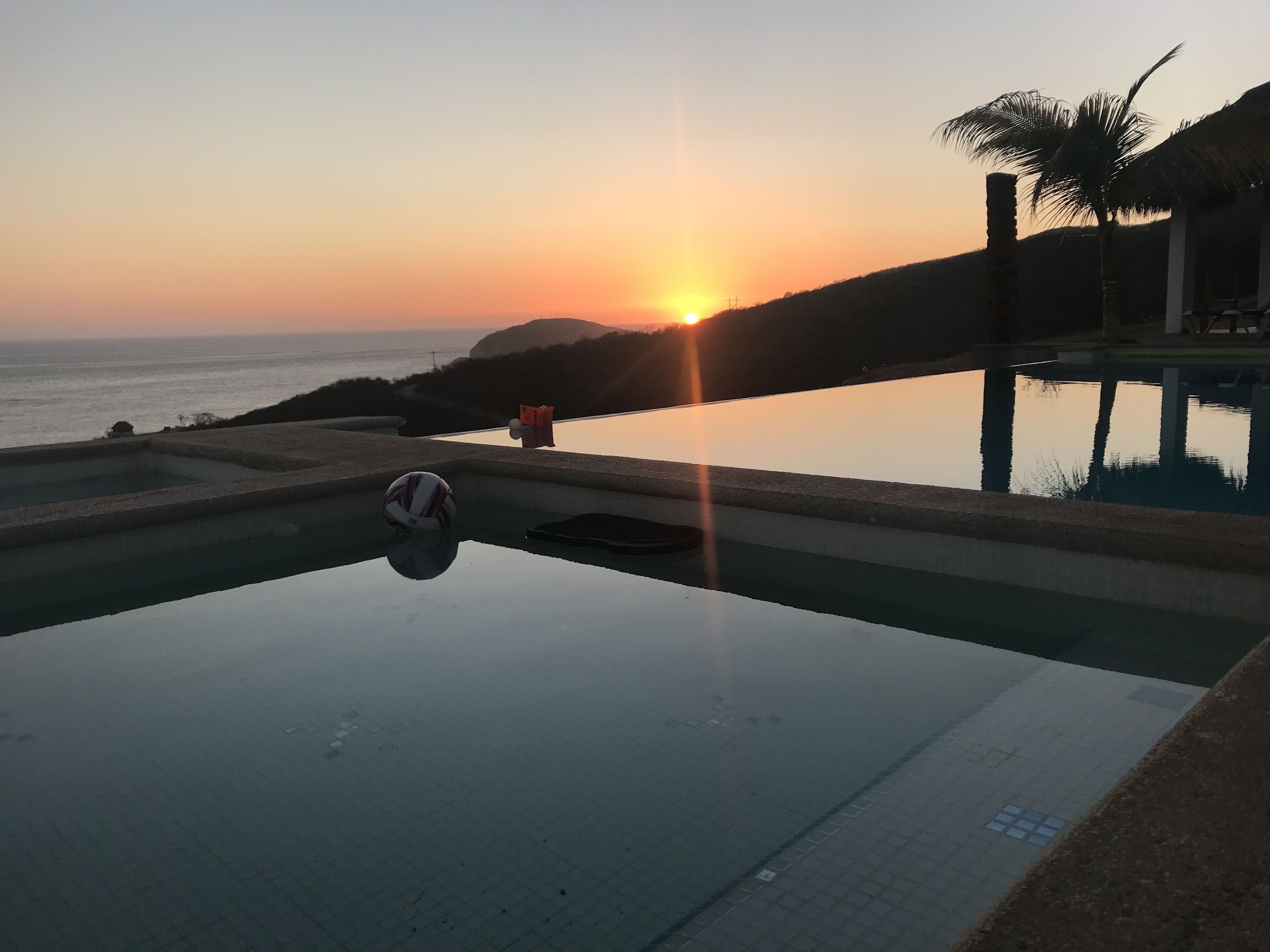 Una piscina con una voleibol, una palmera y una puesta de sol al fondo.