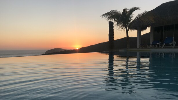 Un atardecer tranquilo sobre una piscina serena, con palmeras y sillas de descanso.