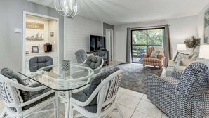 A modern living room with a glass dining table, white wicker chairs, a television, and a patterned sofa. - Central Oasis: Pool, Tennis, Screened Porch Gem! (Fernandina Beach)