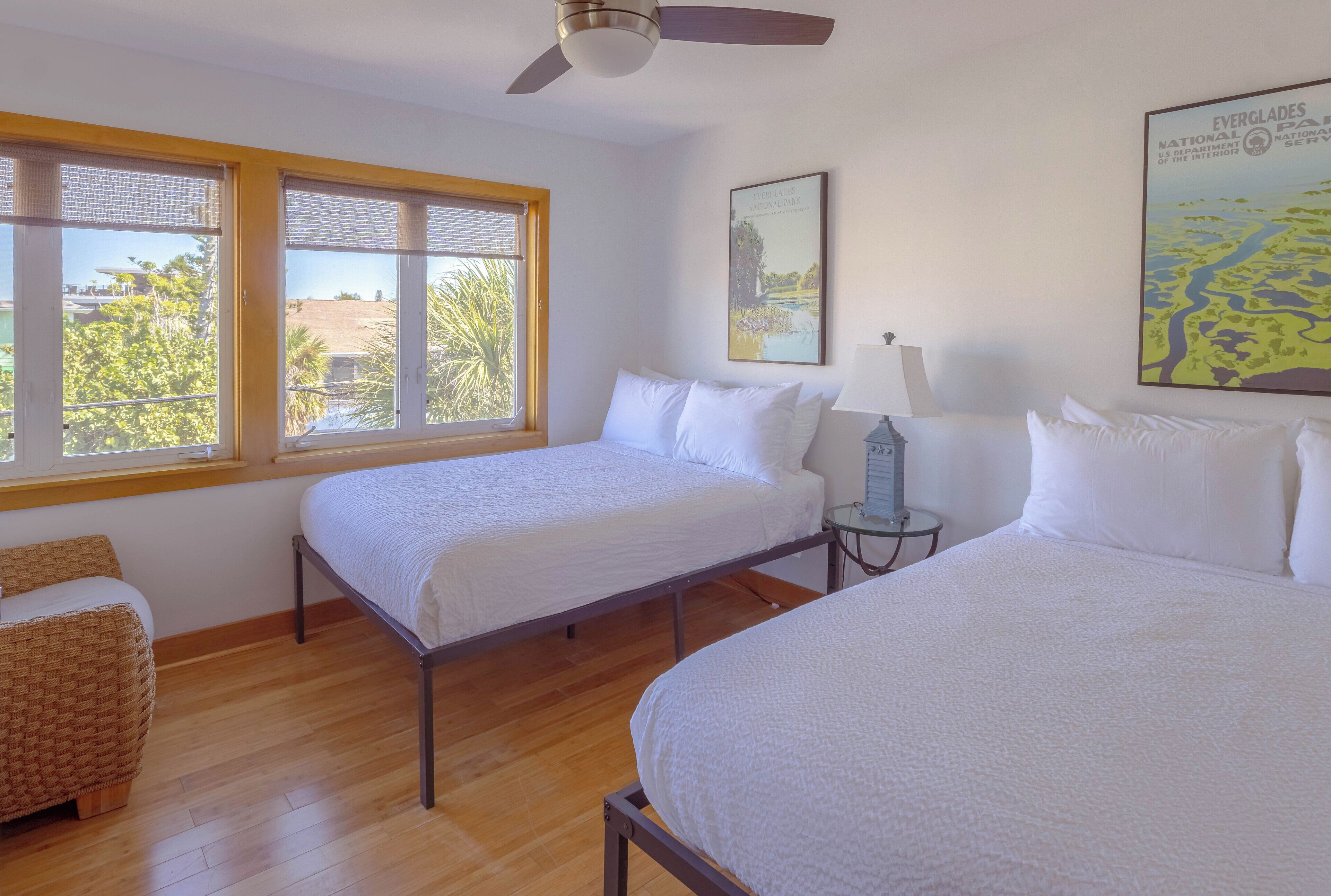 A hotel room with two beds, a ceiling fan, a window with blinds, a framed picture on the wall, and a small table with a lamp.