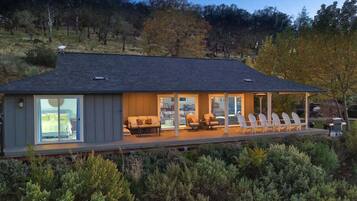 A modern house with a large glass window, a covered porch with chairs, and a view of trees.