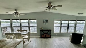 A room with a dining table and chairs, a ceiling fan, and large windows overlooking a body of water.
