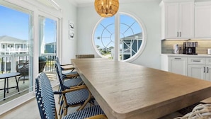 A modern kitchen with white cabinets, a wooden dining table, wicker chairs, and a large window. - Beach Please! (Ocean Isle Beach)