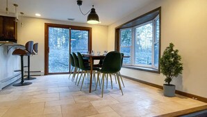 Uma área de refeições com mesa e cadeiras de madeira, uma luminária pendente e vista para árvores através de grandes janelas.