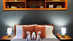 Quarto com cama, duas luminárias de cabeceira, mesa de cabeceira, prateleira de madeira sobre a cama e uma pequena planta em vaso.