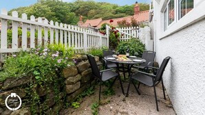 Une terrasse avec une table ronde et des chaises, entourée d’une clôture blanche en bois et d’une végétation luxuriante.
