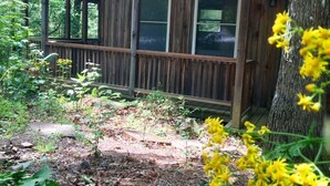 A wooden cabin with a porch, labeled "Bear Den," surrounded by natural vegetation and a tree. - Cabins at Brush Creek Mtn-Bear Den (Del Rio)