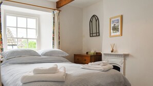 A neatly made bed with striped bedding and folded towels, a wooden nightstand with a tissue box, a fireplace with a framed picture above it, and a window with floral curtains. - 3 bedroom accommodation in Boscastle (Boscastle)