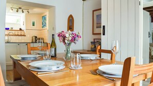 A dining area with a wooden table set for four, surrounded by chairs. A vase with flowers is on the table, and there are wine glasses and plates. The room has white walls and wooden beams on the ceiling. - 3 bedroom accommodation in Boscastle (Boscastle)