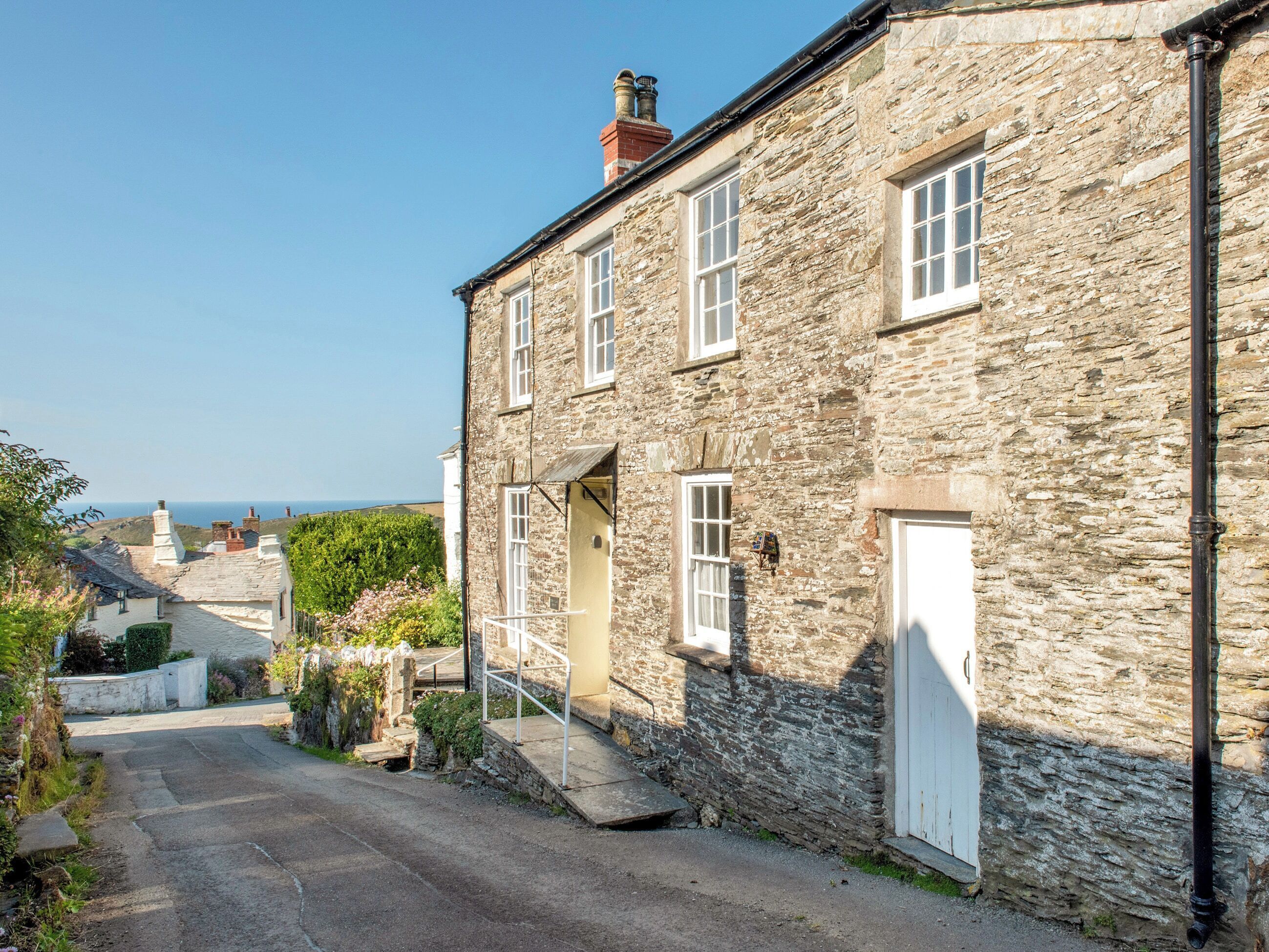 A stone building with a white door and windows, a narrow street, and a view of the sea.
