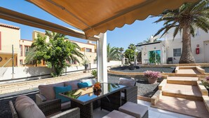 Uma área de estar externa com mobiliário de vime, uma mesa com tampo de vidro e vista para os edifícios e palmeiras ao redor.