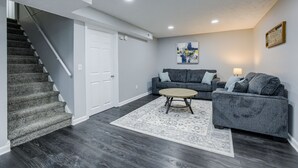 A modern living room with a grey sofa set, a coffee table, and a patterned rug. A staircase with dark wooden steps leads to a white door. - Hamilton in heart of Omaha! Multi family and group friendly! The perfect space!! (Omaha)